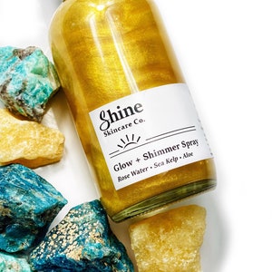 May include: A clear glass bottle filled with gold shimmer spray, labeled "Shine Skincare Co." with "Glow + Shimmer Spray." The bottle is surrounded by turquoise and yellow rough-cut stones, creating a spa-like aesthetic.
