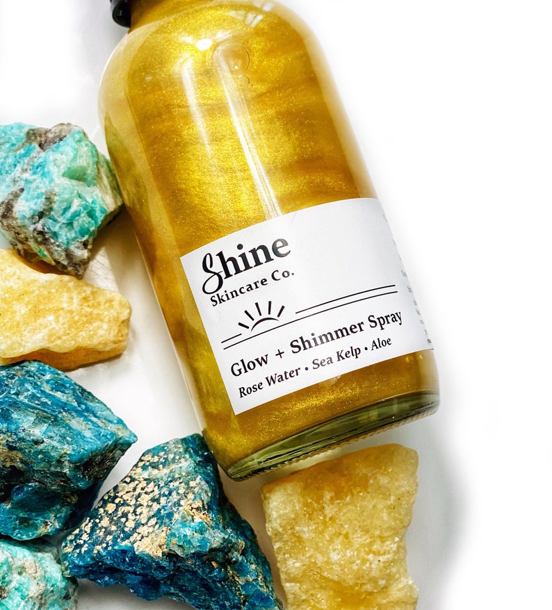May include: A clear glass bottle filled with gold shimmer spray, labeled "Shine Skincare Co." with "Glow + Shimmer Spray." The bottle is surrounded by turquoise and yellow rough-cut stones, creating a spa-like aesthetic.
