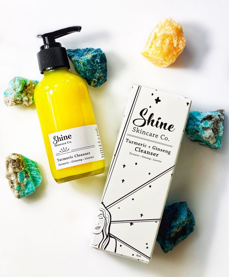 Organic skincare turmeric cleanser in clear glass bottle with pump dispenser. Face wash is bright yellow color and photographed on white background with white shine skincare box. Skincare products surrounded by blue and yellow crystals.