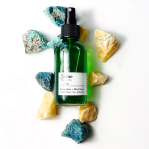 May include: A green glass bottle with a black spray top and a white label that reads "Shine Skincare Co. Cucumber + Mint Toner Colloidal Silver - Aloe - Willowherb". The bottle is surrounded by small blue and yellow stones.