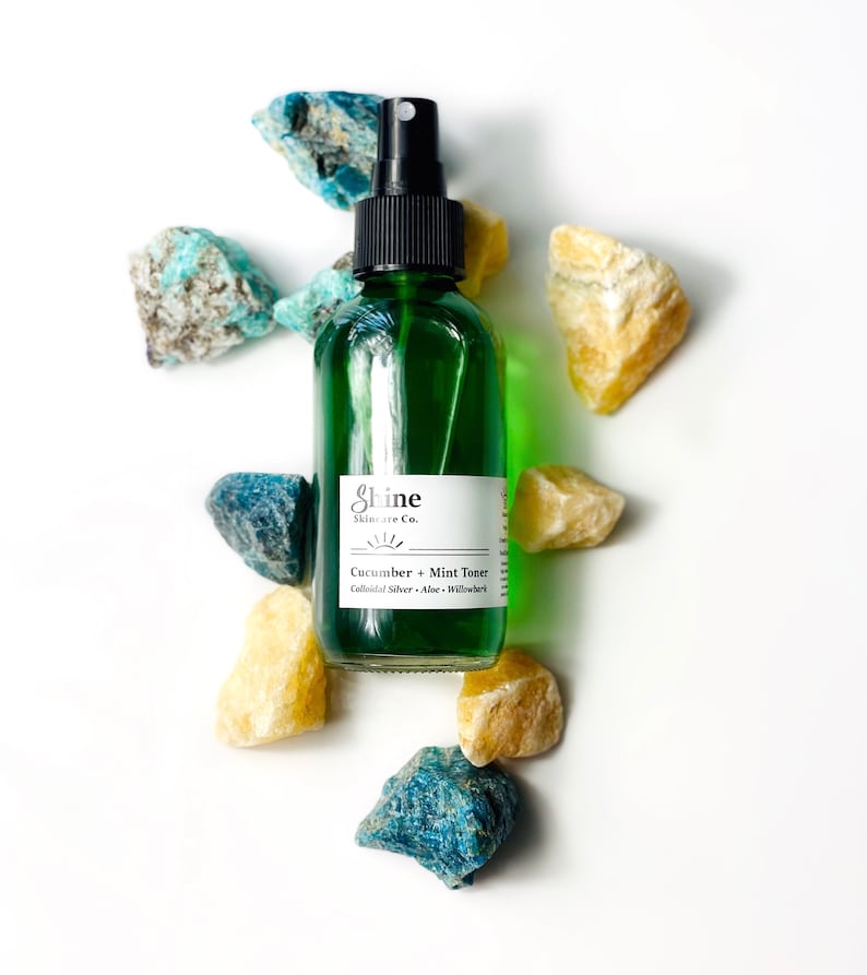 May include: A green glass bottle with a black spray top and a white label that reads "Shine Skincare Co. Cucumber + Mint Toner Colloidal Silver - Aloe - Willowherb". The bottle is surrounded by small blue and yellow stones.