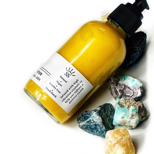 Puede incluir: Una botella de suero amarilla con una bomba negra, rodeada de cristales coloridos. La etiqueta dice "Natural, Vegan, Cruelty Free, Small Batch." El texto en la etiqueta dice "Infused with high vibration crystals to help you radiate a positive GLOW."