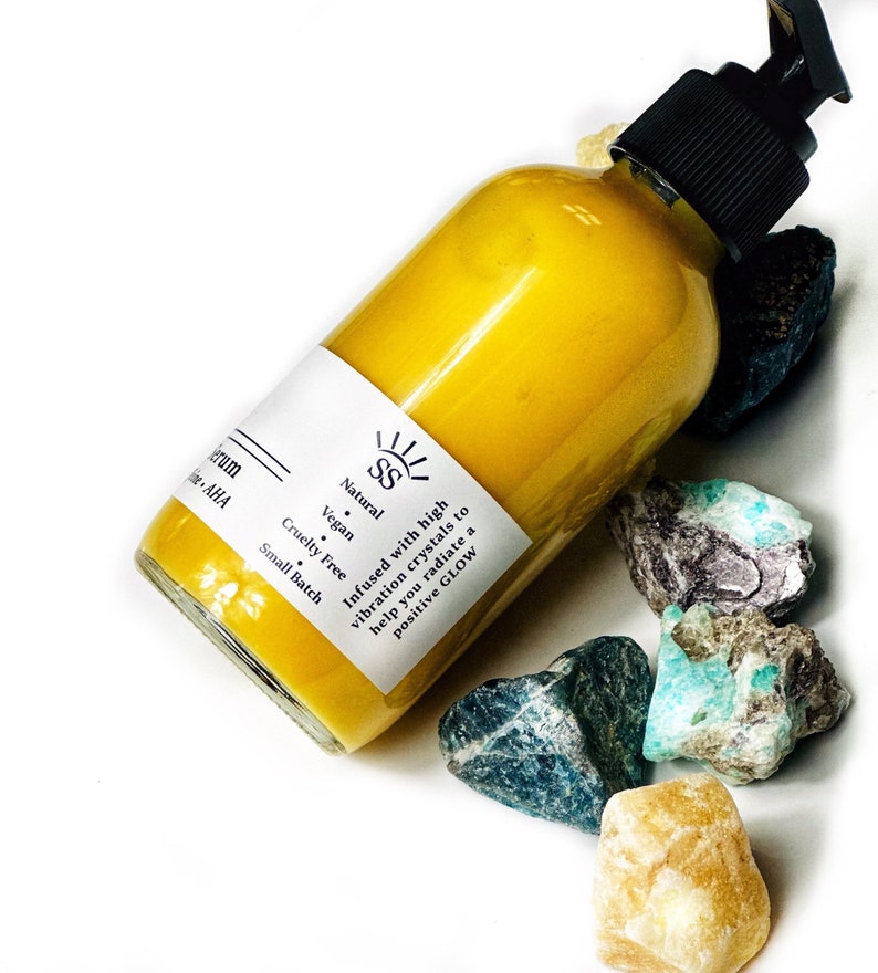 Puede incluir: Una botella de suero amarilla con una bomba negra, rodeada de cristales coloridos. La etiqueta dice "Natural, Vegan, Cruelty Free, Small Batch." El texto en la etiqueta dice "Infused with high vibration crystals to help you radiate a positive GLOW."