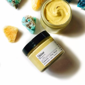 May include: Two glass jars of turmeric clay face mask with a yellow and white label that reads "Shine Skincare Co. Turmeric + Clay Mask Willowbark, Aloe, Turmeric". The jars are on a white surface with several small blue and yellow crystals.