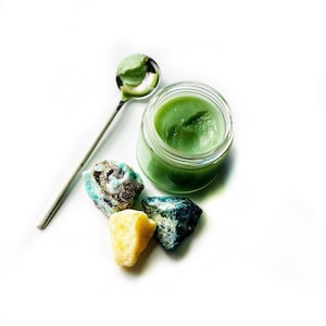 May include: A small glass jar filled with a green balm or cream, surrounded by three small green, blue, and yellow stones. A silver spoon is lying on the white surface.