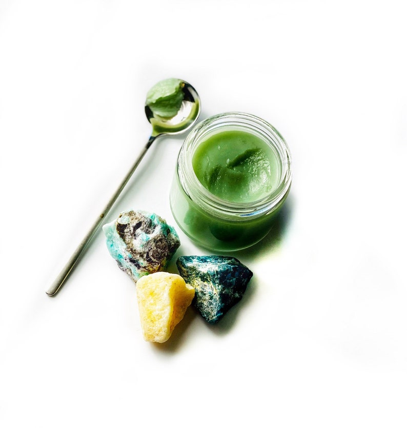 May include: A small glass jar filled with a green balm or cream, surrounded by three small green, blue, and yellow stones. A silver spoon is lying on the white surface.
