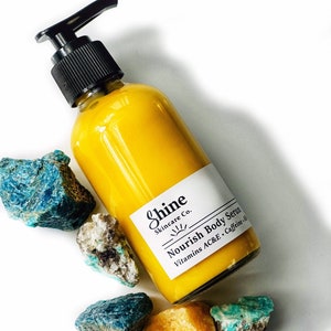 Puede incluir: Una botella de suero corporal amarillo con una bomba negra, etiquetada como "Shine Skincare Co. Nourish Body Serum". La botella est&aacute; rodeada de rocas azules, verdes y amarillo-naranja. El suero contiene vitaminas A, C, E y cafe&iacute;na.