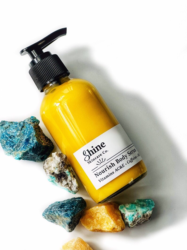 Puede incluir: Una botella de suero corporal amarillo con una bomba negra, etiquetada como "Shine Skincare Co. Nourish Body Serum". La botella est&aacute; rodeada de rocas azules, verdes y amarillo-naranja. El suero contiene vitaminas A, C, E y cafe&iacute;na.