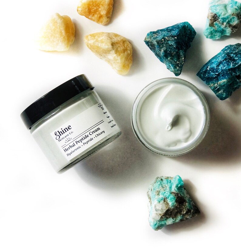 Organic skincare anti-aging face moisturizer in clear glass jar with black lid. Face cream is a very pale blue color. Photo of two jars on white background with blue and yellow crystal gemstones. One jar is open to show cream from top.