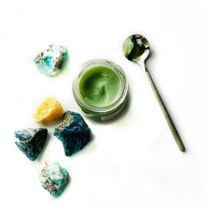 May include: A small jar of green balm with a silver spoon and several blue and green crystals on a white background.