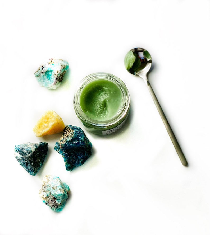 May include: A small jar of green balm with a silver spoon and several blue and green crystals on a white background.