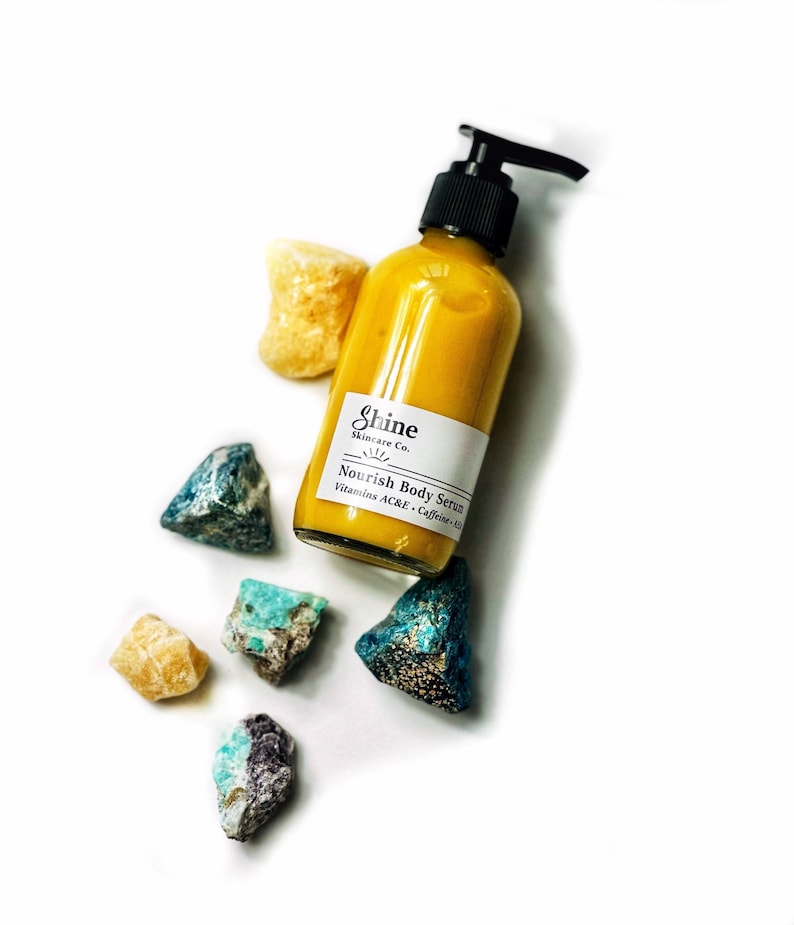 Puede incluir: Una botella de vidrio transparente de Shine Skincare Co. Nourish Body Serum con un dispensador de bomba negro. El suero amarillo est&aacute; rodeado de piedras preciosas de colores y en bruto sobre un fondo blanco.
