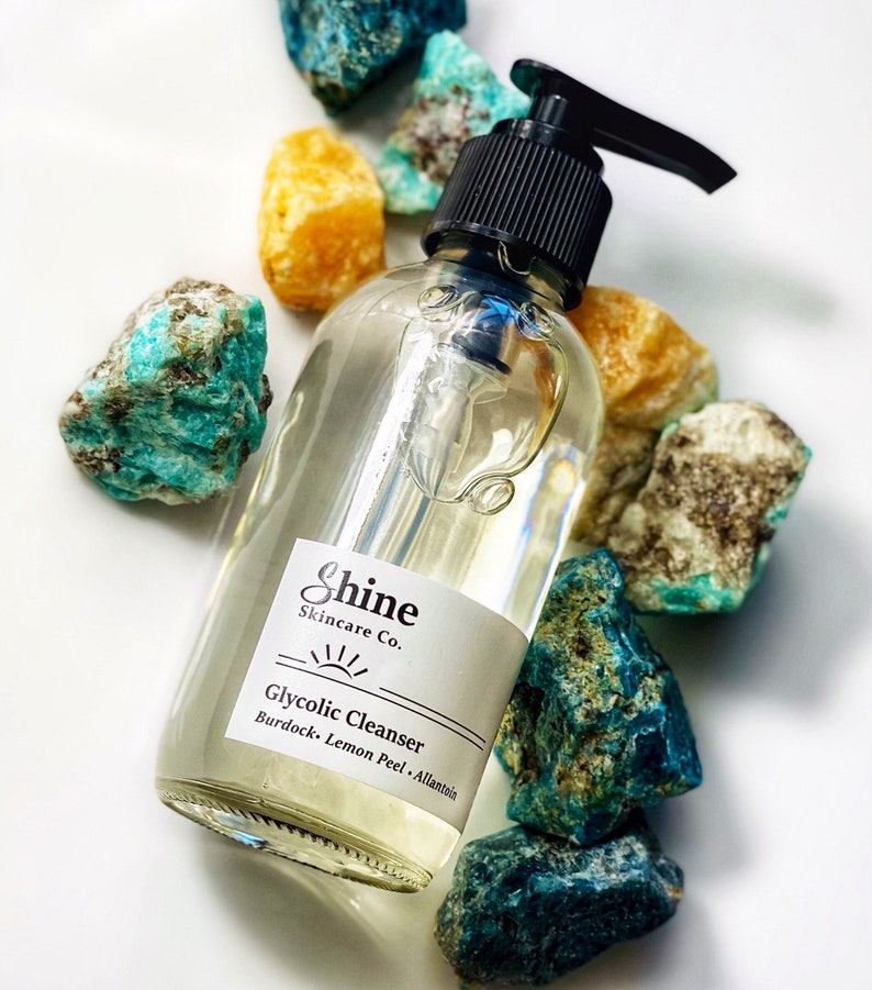 Pu&ograve; includere: Una bottiglia di vetro trasparente con un tappo a pompa nero e un'etichetta bianca che recita "Shine Skincare Co. Glycolic Cleanser Burdock, Lemon Peel, Allantoin". La bottiglia &egrave; circondata da pietre blu e gialle.
