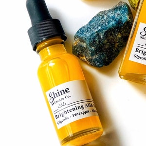 May include: A yellow glass bottle with a black dropper top. The bottle is labelled "Shine Skincare Co. Brightening AHA Serum Glycolic, Pineapple, Niacinamide".