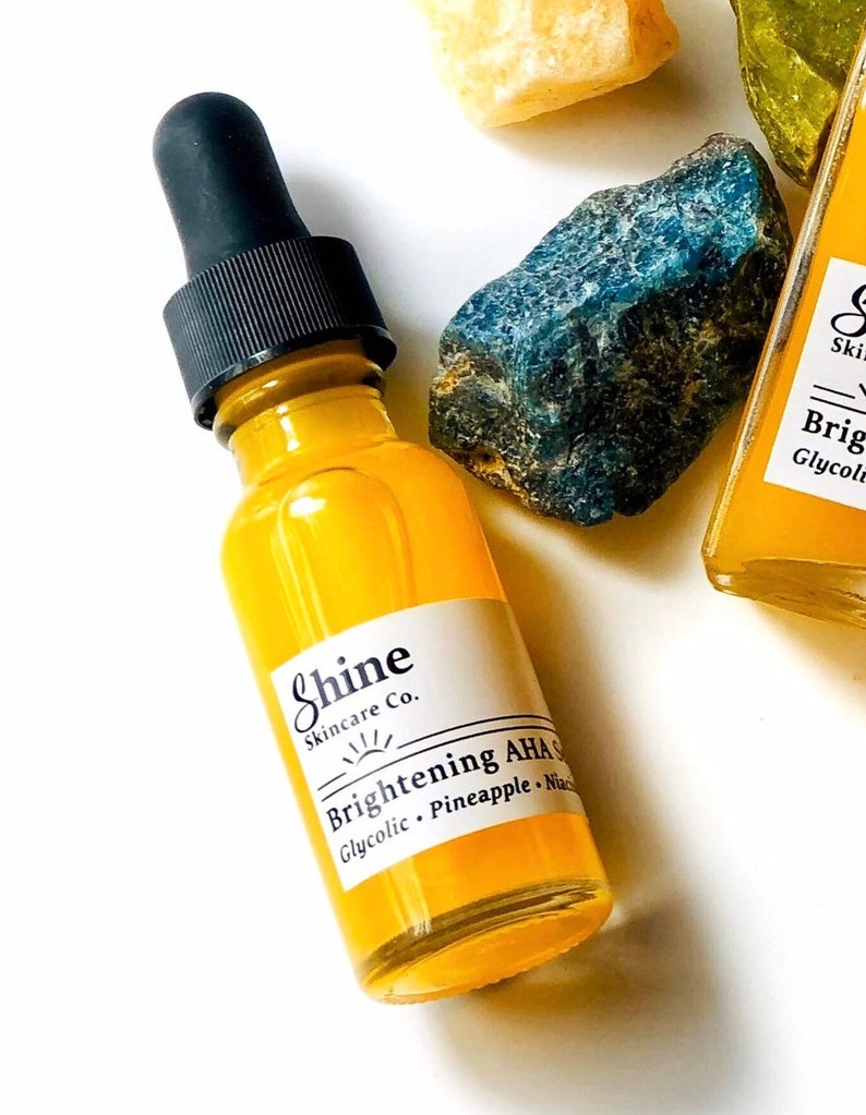May include: A yellow glass bottle with a black dropper top. The bottle is labelled "Shine Skincare Co. Brightening AHA Serum Glycolic, Pineapple, Niacinamide".