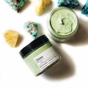 Organic skincare superfood clay face mask is packaged in a clear glass jar. Clay mask is a light green color with a black lid. Skincare product is photographed on a white background with yellow and blue crystal gemstones around the face mask.