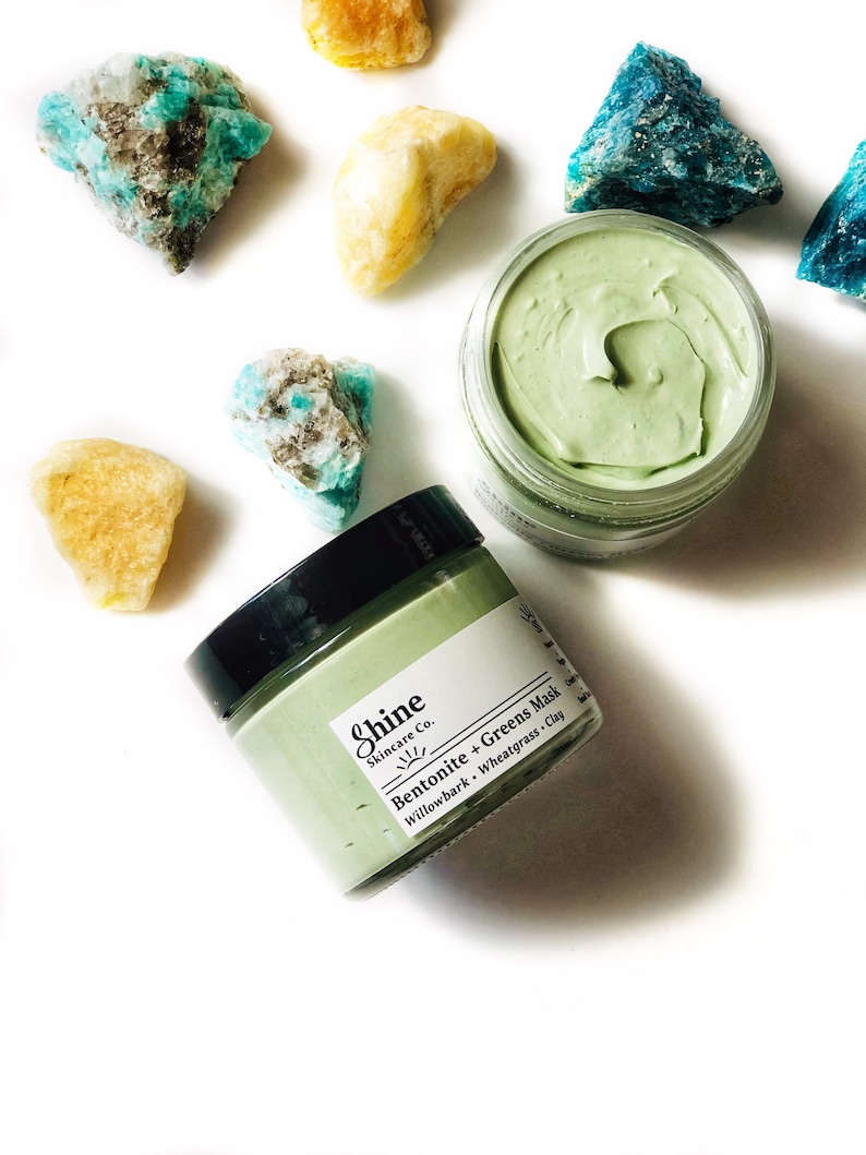 Organic skincare superfood clay face mask is packaged in a clear glass jar. Clay mask is a light green color with a black lid. Skincare product is photographed on a white background with yellow and blue crystal gemstones around the face mask.