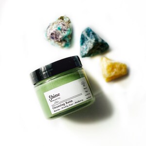 May include: A green jar of cleansing balm with the text "Shine Skincare Co. Cleansing Balm Matcha - Prickly Pear - Mulberry" on the label. The jar is sitting on a white surface with three colorful stones.