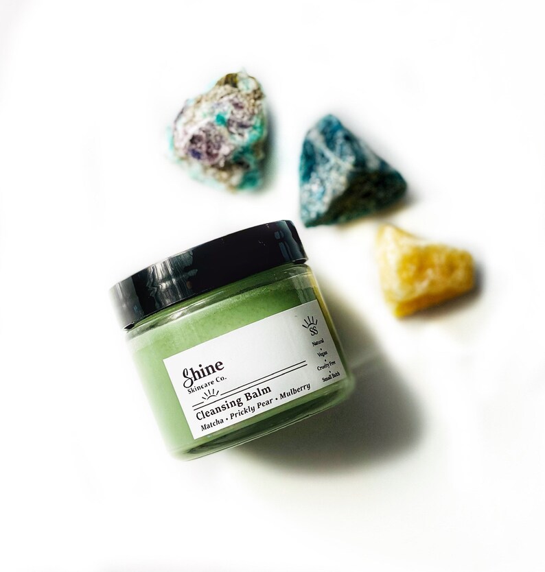May include: A green jar of cleansing balm with the text "Shine Skincare Co. Cleansing Balm Matcha - Prickly Pear - Mulberry" on the label. The jar is sitting on a white surface with three colorful stones.
