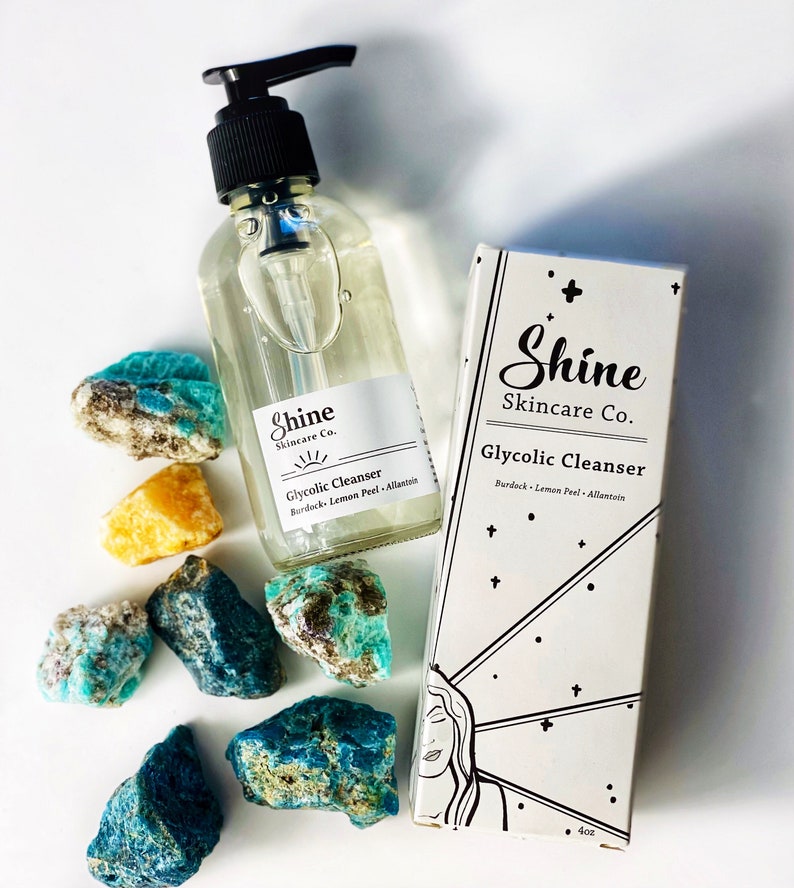 Pu&ograve; includere: Un flacone di vetro trasparente con un erogatore a pompa nero riempito di un liquido trasparente. Il flacone &egrave; etichettato "Shine Skincare Co. Glycolic Cleanser Burdock Lemon Peel Allantoin".  Accanto al flacone c'&egrave; una scatola bianca con testo nero che recita "Shine Skincare Co. Glycolic Cleanser Burdock Lemon Peel Allantoin" e un'illustrazione in bianco e nero di un viso femminile. Il flacone e la scatola sono circondati da cristalli blu e gialli.