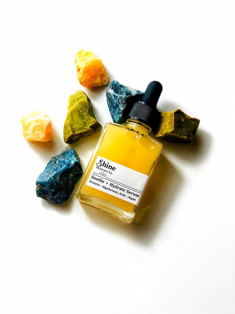 May include: A clear glass bottle of serum with a black dropper lid. The bottle is labeled "Shine Skincare Co. Soothe + Hydrate Serum Protein - Hyaluronic Acid - Algae". The bottle is surrounded by yellow, green, and blue crystals.