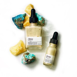 May include: Two glass bottles of facial serum with dropper tops. Both bottles have a white label with the text "Shine Skincare Co." and "Radiance Serum". The larger bottle has the text "Niacinamide Hyaluronic Vitamin E" on the label. The bottles are surrounded by yellow and blue-green stones.