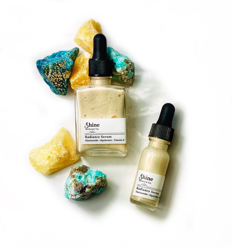 May include: Two glass bottles of facial serum with dropper tops. Both bottles have a white label with the text "Shine Skincare Co." and "Radiance Serum". The larger bottle has the text "Niacinamide Hyaluronic Vitamin E" on the label. The bottles are surrounded by yellow and blue-green stones.