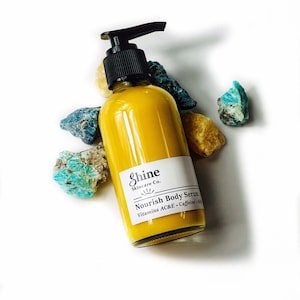 Puede incluir: Una botella de vidrio transparente con un dispensador de bomba negro llena de suero corporal amarillo. La etiqueta dice "Shine Skincare Co. Nourish Body Serum Vitamins AC&E + Caffeine + K2". La botella est&aacute; rodeada de rocas coloridas.