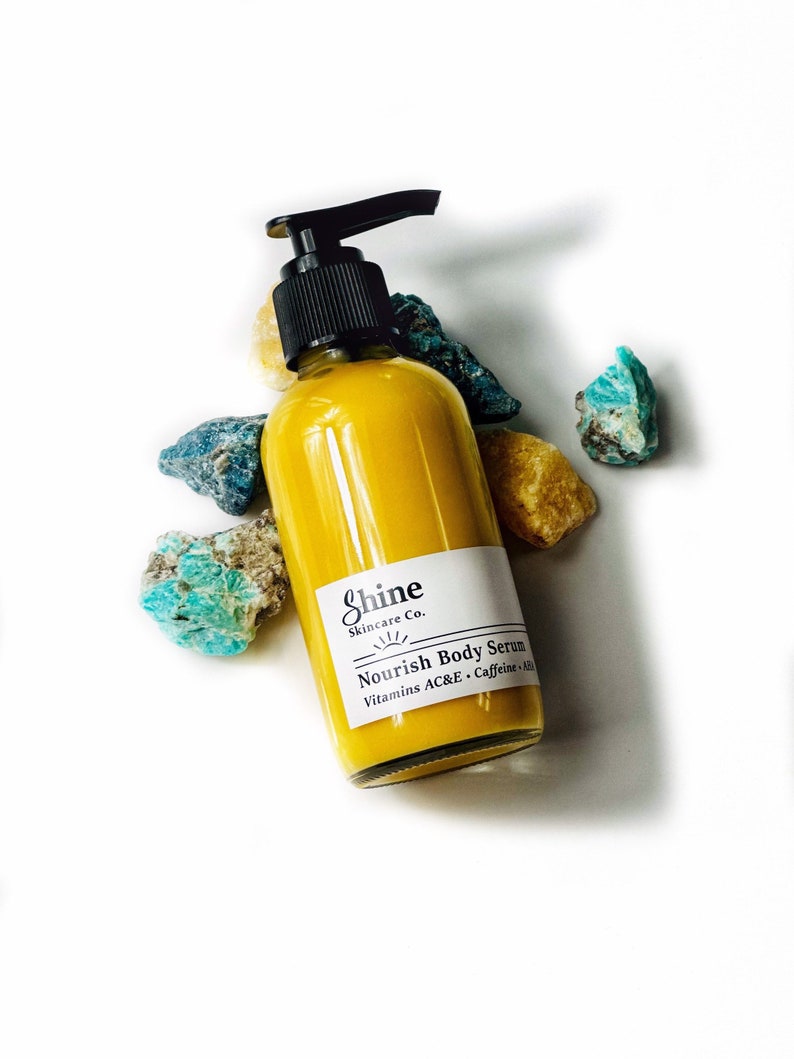 Puede incluir: Una botella de vidrio transparente con un dispensador de bomba negro llena de suero corporal amarillo. La etiqueta dice "Shine Skincare Co. Nourish Body Serum Vitamins AC&E + Caffeine + K2". La botella est&aacute; rodeada de rocas coloridas.