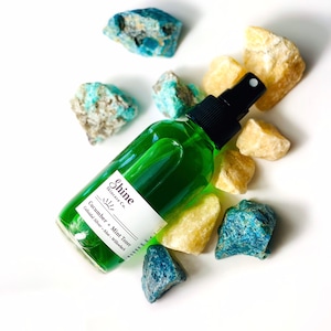Organic skincare cucumber toner in clear glass bottle with black spray top. Face toner is bright green in color. Photo taken on white background with blue and yellow crystal gemstones along the side of the product.