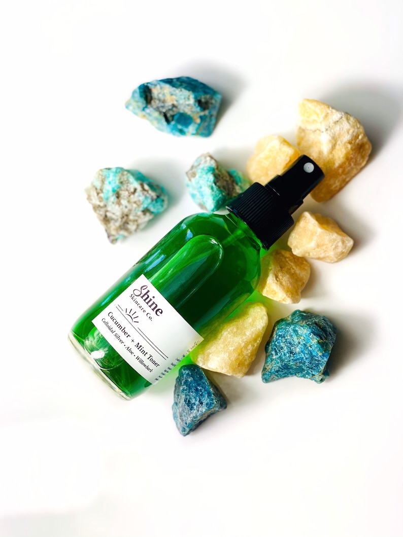 Organic skincare cucumber toner in clear glass bottle with black spray top. Face toner is bright green in color. Photo taken on white background with blue and yellow crystal gemstones along the side of the product.