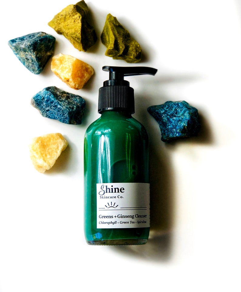 Natural skincare green tea cleanser in clear glass bottle with pump lid. Face wash is a dark green color and thick consistency. White background with blue, green and yellow crystal gemstones around top of bottle.