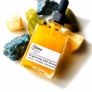 May include: A glass bottle of yellow serum with a black dropper lid. The bottle is labelled "Shine Skincare Co. Brightening AHA Serum Glycolic - Pineapple - Niacinamide". The bottle is surrounded by small blue, green, and yellow stones.