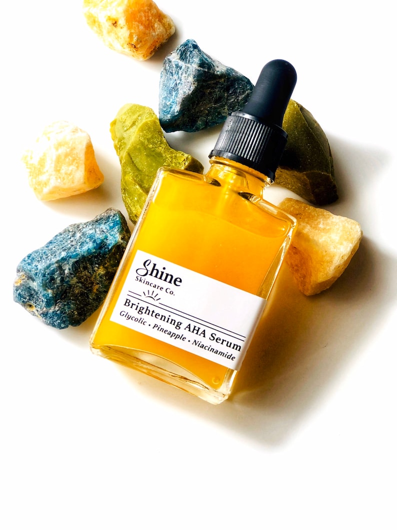 May include: A glass bottle of yellow serum with a black dropper lid. The bottle is labelled "Shine Skincare Co. Brightening AHA Serum Glycolic - Pineapple - Niacinamide". The bottle is surrounded by small blue, green, and yellow stones.