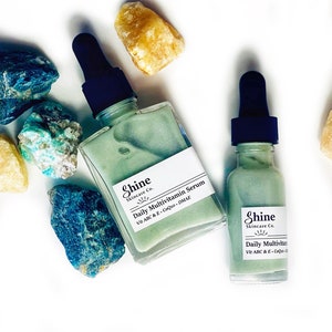 Op de afbeelding: Twee glazen flesjes met groene serum met druppelaars. De flesjes zijn gelabeld met "Shine Skincare Co. Daily Multivitamin Serum Vit ABC & E - CoQ10 - DMAE".