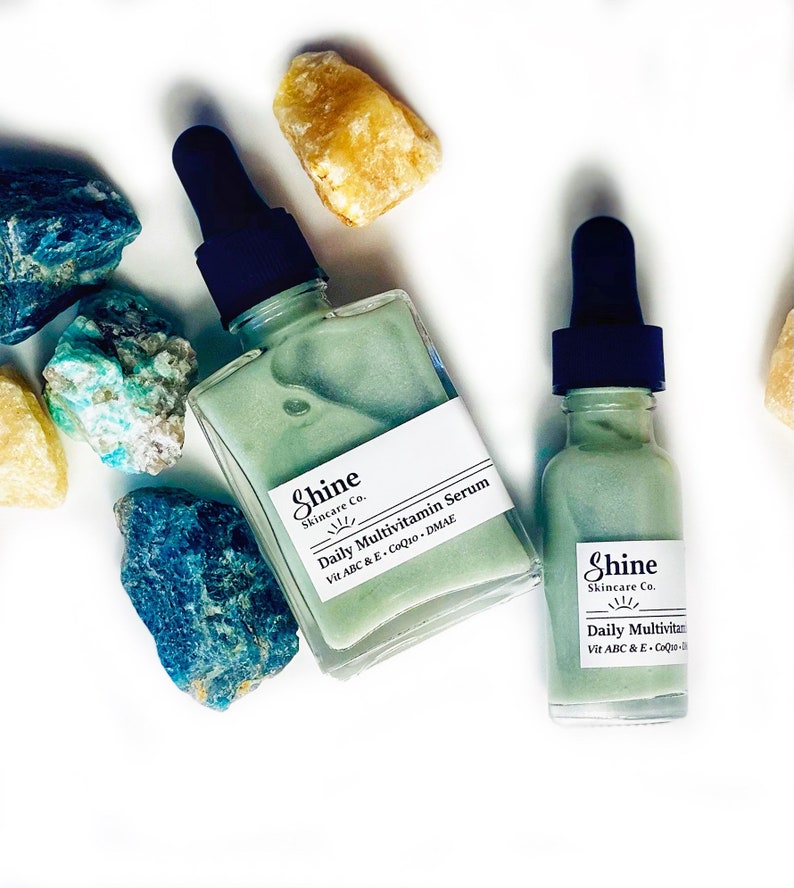 Op de afbeelding: Twee glazen flesjes met groene serum met druppelaars. De flesjes zijn gelabeld met "Shine Skincare Co. Daily Multivitamin Serum Vit ABC & E - CoQ10 - DMAE".