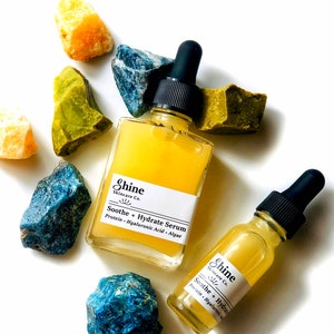 May include: Two glass bottles of serum with dropper tops. The bottles are labeled "Shine Skincare Co. Soothe + Hydrate Serum Protein. Hyaluronic Acid. Algae". The bottles are surrounded by blue and green stones.