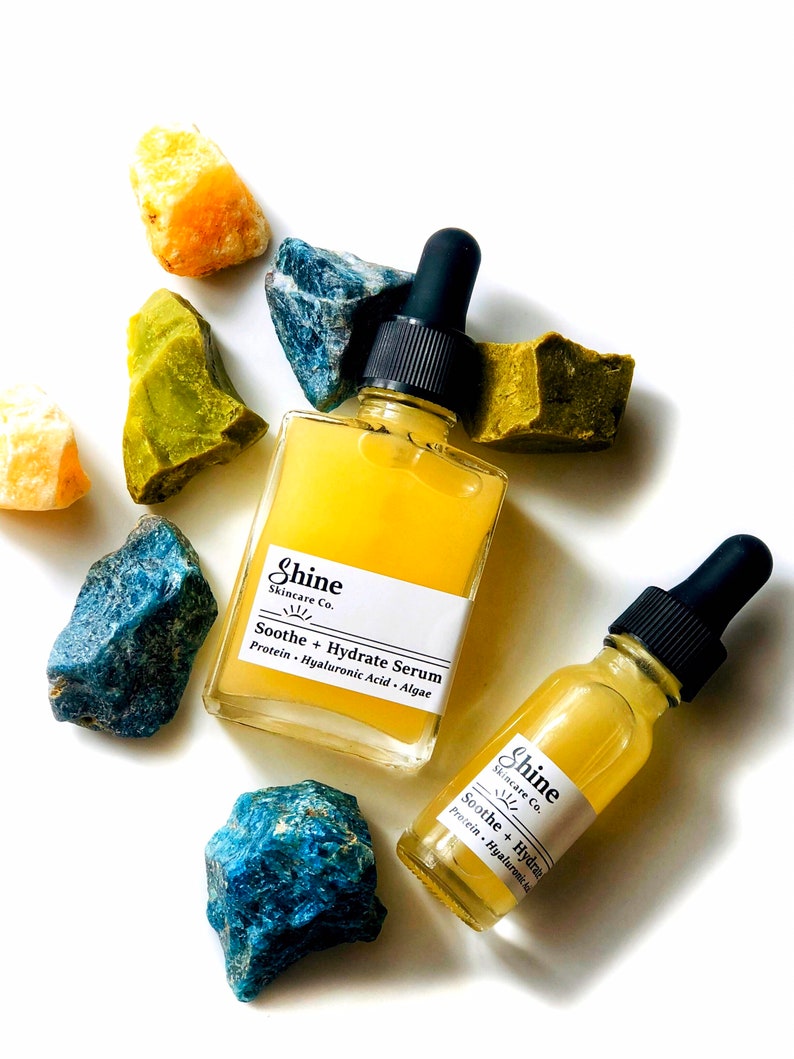 May include: Two glass bottles of serum with dropper tops. The bottles are labeled "Shine Skincare Co. Soothe + Hydrate Serum Protein. Hyaluronic Acid. Algae". The bottles are surrounded by blue and green stones.