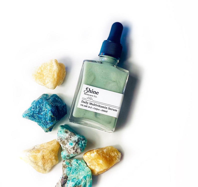 Op de afbeelding: Een groene glazen fles met een druppelaar, met het label "Shine Skincare Co. Daily Multivitamin Serum with A, B, C & E, CoQ10, DMAE". De fles is omringd door gele en blauwe stenen.
