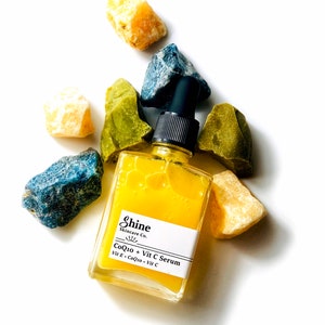 Organic skincare vitamin c serum in clear square glass bottle. Serum is pale yellow color and thin consistency. Photographed on white background with blue, yellow and green crystals surrounding serum.