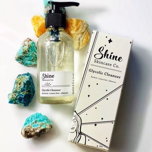Pu&ograve; includere: Una bottiglia di vetro trasparente di detergente glicolico con un tappo a pompa nero. La bottiglia &egrave; etichettata "Shine Skincare Co. Glycolic Cleanser Burdock Lemon Peel Allantoin". La bottiglia &egrave; su una superficie bianca con pietre blu e gialle.