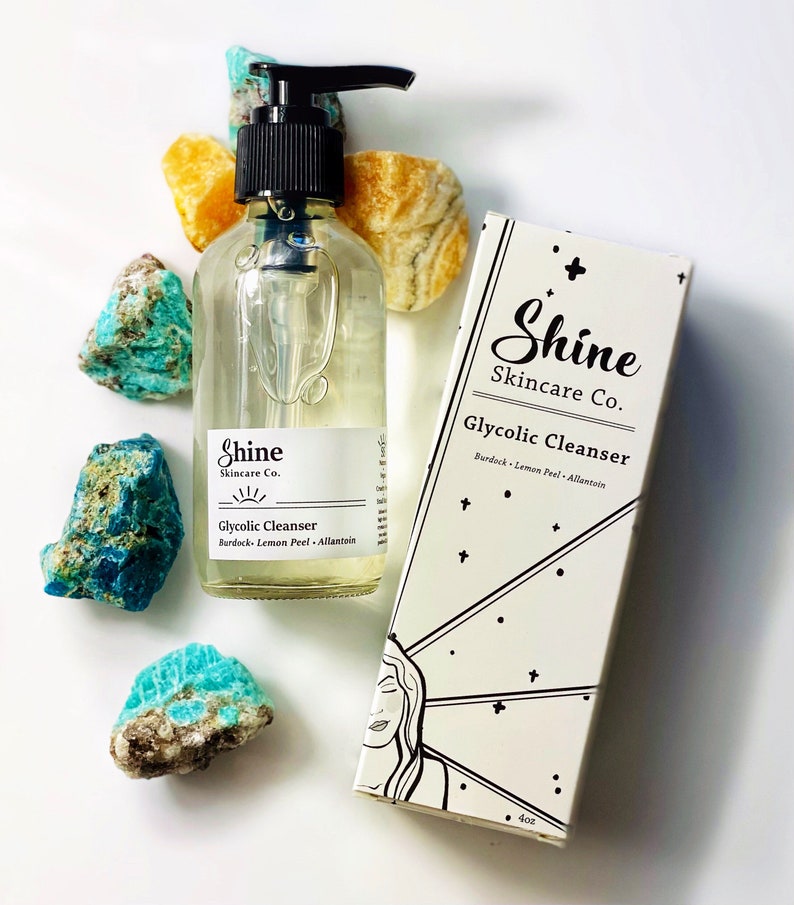 Pu&ograve; includere: Una bottiglia di vetro trasparente di detergente glicolico con un tappo a pompa nero. La bottiglia &egrave; etichettata "Shine Skincare Co. Glycolic Cleanser Burdock Lemon Peel Allantoin". La bottiglia &egrave; su una superficie bianca con pietre blu e gialle.