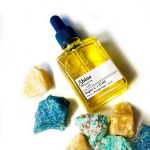 May include: A square glass bottle of yellow oil with a dark blue dropper, labeled "Shine Skincare Co. Super C + E Oil." The bottle is surrounded by various colorful gemstones, including blue, turquoise, and yellow-orange rocks, on a white background.
