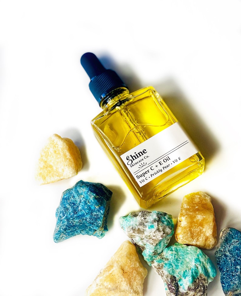 May include: A square glass bottle of yellow oil with a dark blue dropper, labeled "Shine Skincare Co. Super C + E Oil." The bottle is surrounded by various colorful gemstones, including blue, turquoise, and yellow-orange rocks, on a white background.