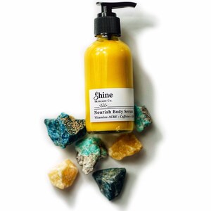 Puede incluir: Una botella de suero corporal amarillo con un dispensador de bomba negro y una etiqueta blanca que dice "Shine Skincare Co." y "Nourish Body Serum". La botella est&aacute; rodeada de piedras preciosas de colores en bruto. El suero contiene Vitaminas AC&E y Cafe&iacute;na.