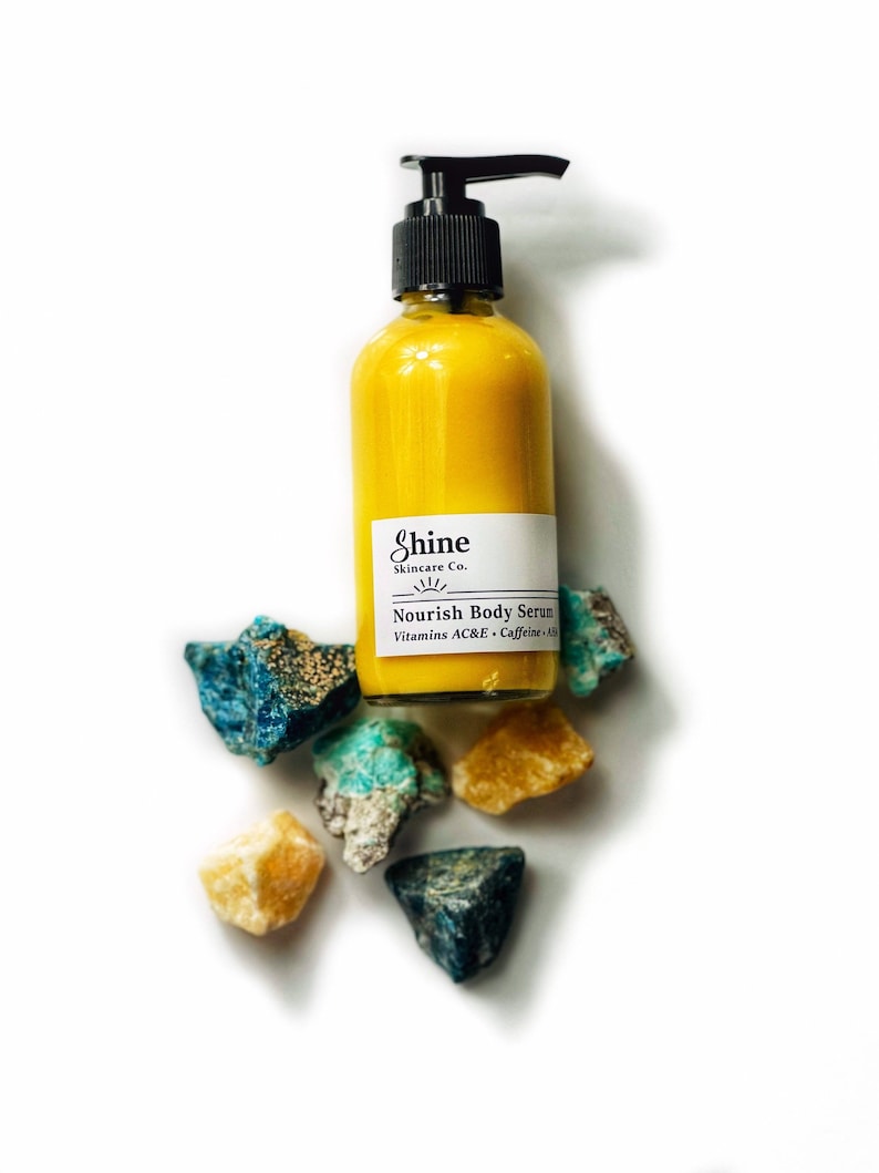 Puede incluir: Una botella de suero corporal amarillo con un dispensador de bomba negro y una etiqueta blanca que dice "Shine Skincare Co." y "Nourish Body Serum". La botella est&aacute; rodeada de piedras preciosas de colores en bruto. El suero contiene Vitaminas AC&E y Cafe&iacute;na.