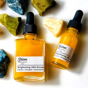 May include: Two glass bottles of yellow serum with dropper tops. The bottles are labeled 'Shine Skincare Co. Brightening AHA Serum Glycolic - Pineapple - Niacinamide
