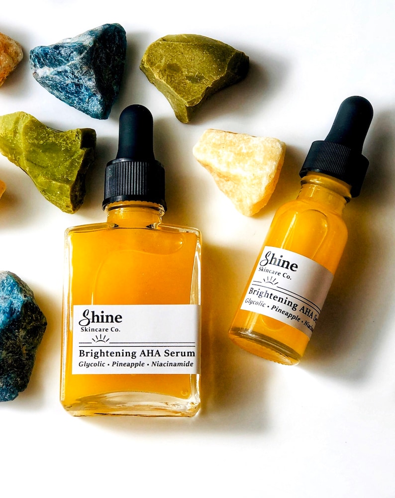 May include: Two glass bottles of yellow serum with dropper tops. The bottles are labeled 'Shine Skincare Co. Brightening AHA Serum Glycolic - Pineapple - Niacinamide