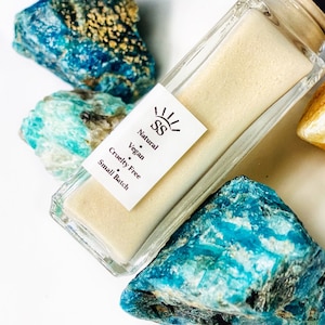 May include: A clear glass bottle with a white label that reads "Natural, Vegan, Cruelty Free, Small Batch" and the initials "SS" in a sunburst design. The bottle is sitting on top of blue and green stones.