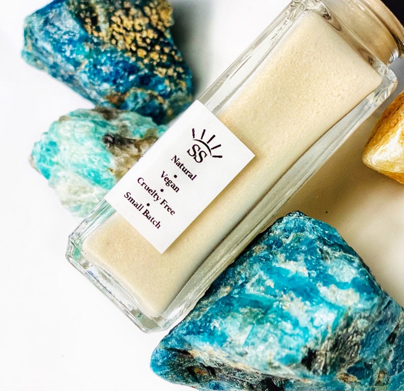 May include: A clear glass bottle with a white label that reads "Natural, Vegan, Cruelty Free, Small Batch" and the initials "SS" in a sunburst design. The bottle is sitting on top of blue and green stones.